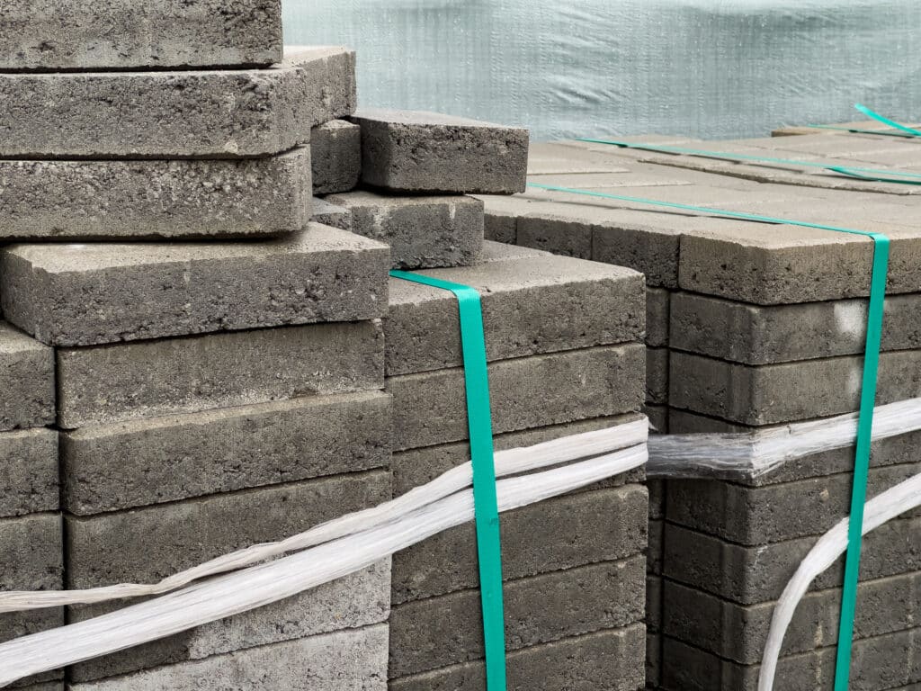 gray brick pavers are neatly stacked and secured with straps, ready for use in paving a sidewalk on an urban street, indicating construction activity nearby